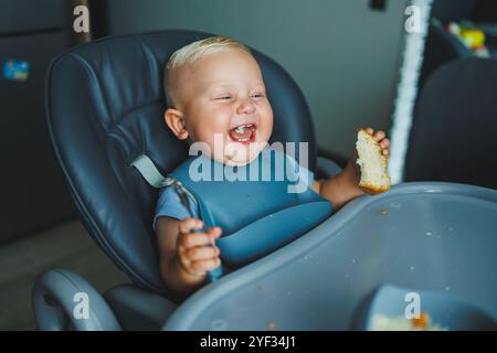 Un garçon de 1 an est assis dans une chaise haute et mange du pain. Le bébé mange du pain. L'auto-alimentation de l'enfant. Banque D'Images