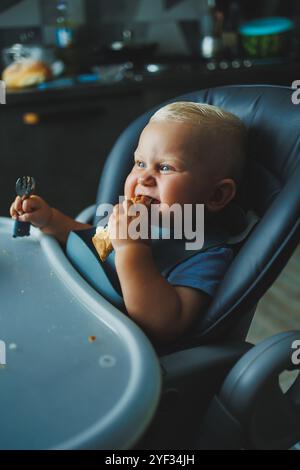 Un garçon de 1 an est assis dans une chaise haute et mange du pain. Le bébé mange du pain. L'auto-alimentation de l'enfant. Banque D'Images