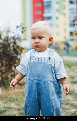 Un petit garçon de 1 an en salopette en denim marche dans la rue. Grandes combinaisons pour enfants. Petit garçon élégant sur le terrain de jeu. Banque D'Images