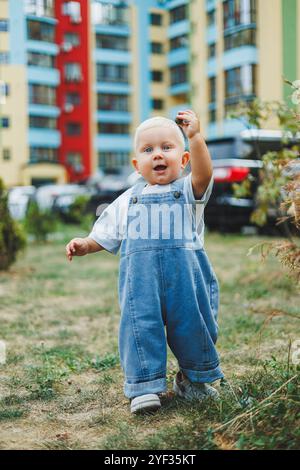 Un petit garçon de 1 an en salopette en denim marche dans la rue. Grandes combinaisons pour enfants. Petit garçon élégant sur le terrain de jeu. Banque D'Images