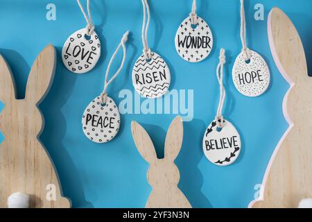 Groupe d'oreilles de lapin en bois et d'œufs de Pâques en argile avec des mots RISEN HOPE BELIEVE PEACE LOVE WONDER faites-le vous-même sécher l'air de l'artisanat d'argile pour les vacances de Pâques. Décoration idée cadeau artisanal pour enfants fait à la main. Instruction étape par étape de la peinture, pressant des chiffres sur fond bleu Banque D'Images