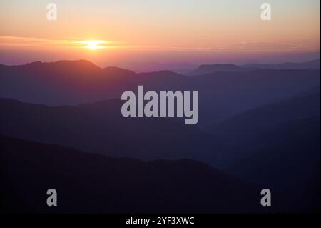 Le soleil se couche sur les montagnes, projetant une lueur dorée chaude à travers le ciel. Les couches de collines s'estompent dans des nuances douces de violet et de bleu. Ambiance paisible de coucher de soleil met en valeur la beauté naturelle du paysage. Banque D'Images