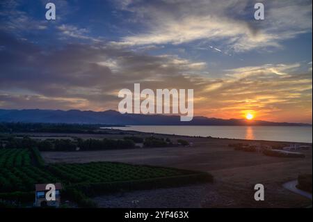 Des rayons dorés de coucher de soleil illuminent un vignoble paisible au bord du lac, encadré par des montagnes lointaines sous un ciel coloré. Banque D'Images