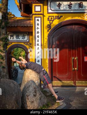 Un homme se penche en arrière sur un rocher pendant qu'il fait des exercices matinaux à l'extérieur de l'ancienne pagode Tran Quoc sur le lac de l'Ouest, Hanoi, Vietnam. Banque D'Images