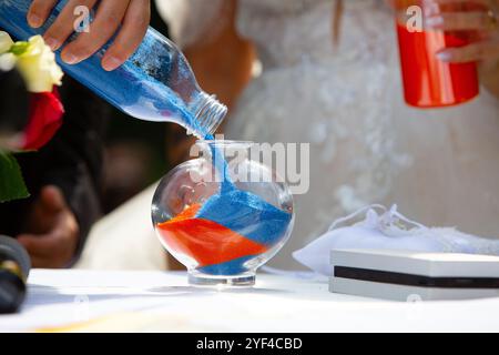 Mariée et mariée effectuant le rituel de cérémonie de sable, versant du sable bleu et orange dans un vase en verre pendant leur cérémonie de mariage Banque D'Images