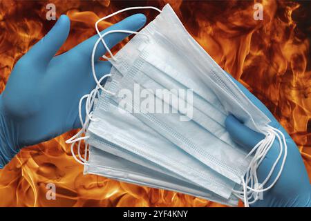 Mains humaines tenant des masques de protection lot dans des gants médicaux bleus sur fond de flammes de feu rouge fort. Concept quarantaine, grippe, pandémie Banque D'Images