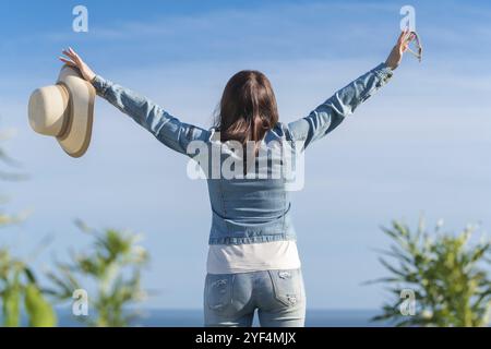 Vue arrière de Happiness hipster femelle a levé les mains profitant des vacances, debout sur fond de ciel bleu sans nuages. Femme méconnaissable habillée en de Banque D'Images