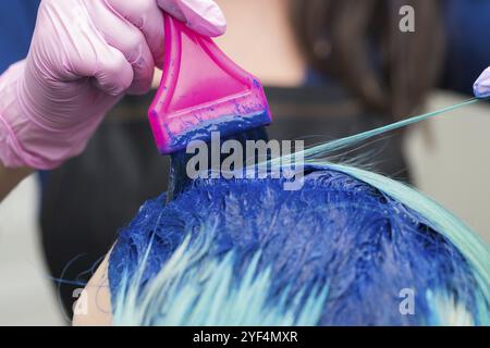 Coiffeurs professionnels en gant de protection à l'aide de la brosse rose tout en appliquant la peinture bleue à la femme avec la couleur de cheveux émeraude, pendant le processus de teinture h Banque D'Images