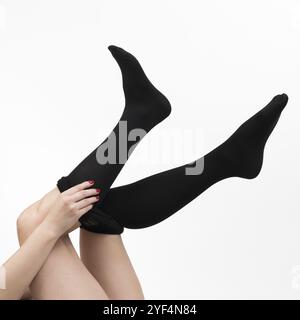 Vue en gros plan des mains de femme habillant les collants noirs sur ses jambes soigneusement. Vue recadrée de jambes féminines méconnaissables sur fond blanc. Prise de vue en studio Banque D'Images