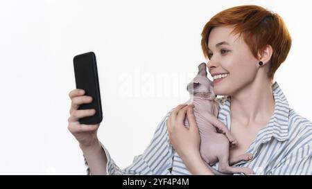 Souriant rousse jeune femme embrassant Sphynx Cat, prend photo selfie sur smartphone, regardant la caméra du téléphone portable. Magnifique hipster habillé de rayures Banque D'Images