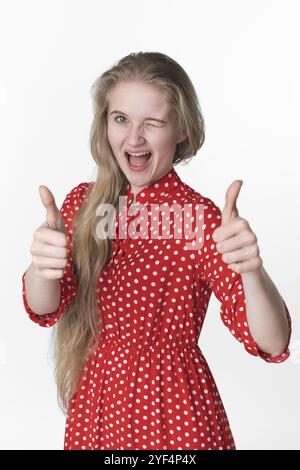 Bonheur blonde jeune femme ludique fermé un œil et montrant les pouces vers le haut avec les deux mains. Portrait de jeune femme adulte souriante habillée en été Banque D'Images