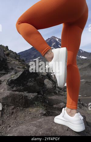 Femme se tient sur une jambe pendant l'entraînement de fitness dans la zone montagneuse. Jambes féminines athlétiques portant des leggings d'entraînement orange et des baskets. Gros plan de la jambe Banque D'Images