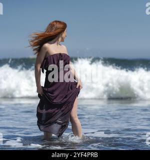 Redhead femme en robe longue avec genou haut ourlet debout cheville profonde dans l'eau sur la plage et la tête tournée, regardant par-dessus l'épaule à la mer en prévision de Banque D'Images
