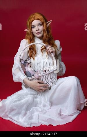 Elfe cosplay jeune femme avec de longs cheveux rouges se trouve sur fond rouge avec un panier en osier blanc sur ses genoux. Il y a drôle de chaton Sphinx dans le panier. Elf In Banque D'Images