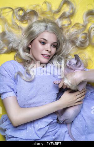 Cosplayer elfe jeune femme blonde en robe bleue d'été couchée sur fond jaune, souriant et embrassant chaton Sphinx. Elfe a luxe bouclé long ha Banque D'Images