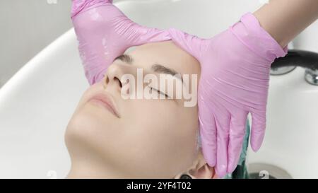 Tête de jeune femme avec les yeux fermés dans l'évier dans le salon de coiffure. Coiffeur mains en gants de protection roses lavage des cheveux dans un salon de beauté professionnel Banque D'Images