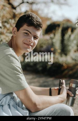 Un adolescent est assis sur un banc dans le parc boit du café dans une tasse thermo et regarde dans un téléphone. Portrait de beau gars joyeux assis sur le banc fres Banque D'Images
