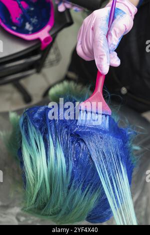 Prise de vue en grand angle de la teinture des cheveux dans un salon de beauté. Coiffer dans un gant de protection à l'aide d'une brosse magenta tout en appliquant de la peinture bleue à la femme avec em Banque D'Images