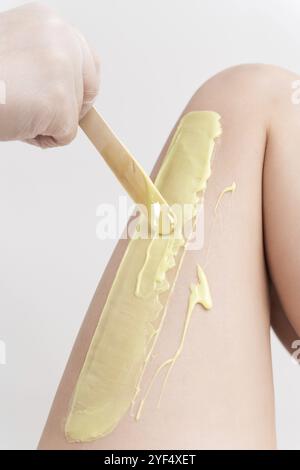 Gros plan main dans le gant en appliquant de la cire chaude verte sur la jambe de la femme à l'aide de la spatule. Procédure professionnelle d'épilation avec de la cire chaude dans le salon de beauté. Partie de la photo Banque D'Images