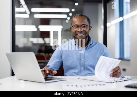 Homme d'affaires afro-américain mature souriant en toute confiance travaillant sur ordinateur portable dans un bureau moderne. Cadre d'affaires avec des documents importants mettant en valeur le succès et le professionnalisme. Banque D'Images