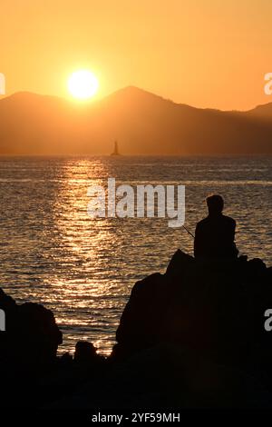 Silhouette de pêcheur assis sur les rochers au coucher du soleil avec vue panoramique sur le massif de l'Estérel depuis le Cap d'Antibes sur la côte d'Azur en grand format Banque D'Images