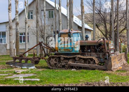 Bulldozer agricole rustique avec une charrue contre une maison de deux étages et une rangée d'arbres Banque D'Images