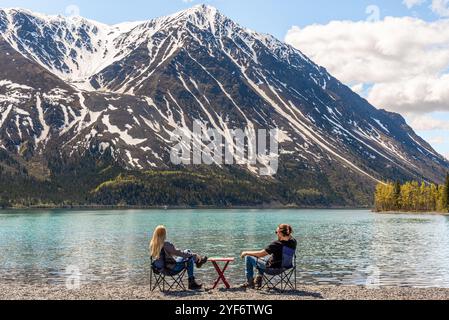 Deux personnes, amis, couple assis sur des chaises de camping avec table de pique-nique sur la zone pittoresque de la rive du lac dans le territoire du Yukon, nord du Canada au printemps Banque D'Images