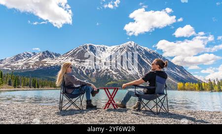 Deux personnes, amis, couple assis sur des chaises de camping avec table de pique-nique sur la zone pittoresque de la rive du lac dans le territoire du Yukon, nord du Canada au printemps Banque D'Images