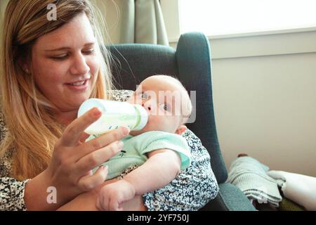 Femme souriante assise dans un fauteuil nourrissant son fils bébé avec une bouteille de lait Banque D'Images
