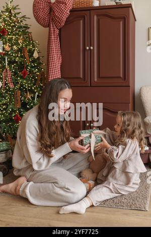 Mère assise sur le sol à côté d'un arbre de Noël illuminé offrant à sa fille un cadeau de Noël emballé Banque D'Images