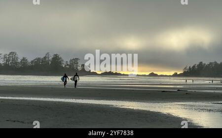 Silhouette de deux surfeurs marchant le long de la plage portant des planches de surf au coucher du soleil, Colombie-Britannique, Canada Banque D'Images