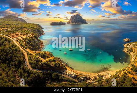 Vue aérienne de la plage de Cala d’Hort à Ibiza, avec le rocher emblématique d’es Vedrà, les eaux turquoises et le littoral méditerranéen pittoresque. Banque D'Images
