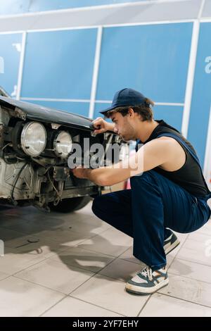 Mécanicien pensif en salopettes accroupissant et se concentrant sur la réparation avant de la voiture endommagée dans un atelier de réparation professionnel. Réparateur évaluant les dommages à la voiture Banque D'Images