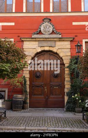 Élégante porte en bois avec des sculptures complexes et une crête décorative sur le bâtiment rouge historique, encadrée par des lanternes vintage, lierre et plantes à fleurs Banque D'Images