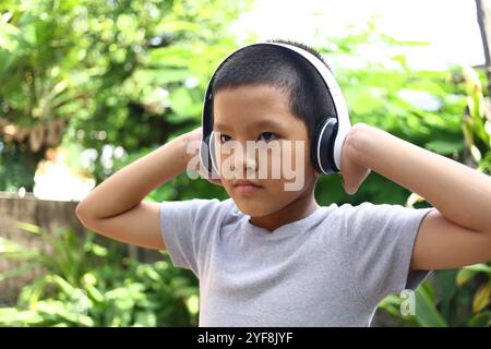 Un jeune garçon écoute de la musique avec des écouteurs, immergé dans un cadre extérieur paisible. La verdure luxuriante rehausse l'atmosphère détendue de la jeunesse et j Banque D'Images