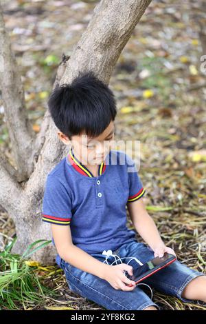 Un jeune garçon est assis sous un arbre, concentré sur son téléphone portable tout en portant des vêtements décontractés. Cette scène extérieure sereine capture le lei contemporain de l'enfance Banque D'Images