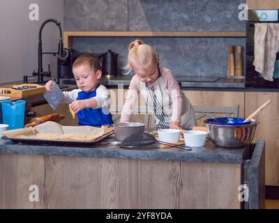 Deux jeunes enfants, frère et sœur, font des biscuits dans la cuisine moderne, plaçant soigneusement la pâte sur des plaques de cuisson que leur travail d'équipe et leur joie font Banque D'Images