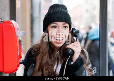 Femme parle au téléphone tout en se tenant debout à côté d'un téléphone public rouge. Elle porte un chapeau noir et une veste noire Banque D'Images