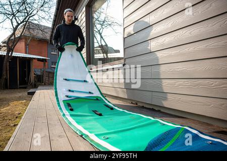 Homme pliant un stand-up paddleboard gonflable (SUP) sur une terrasse en bois à côté d'une maison moderne Banque D'Images