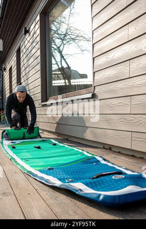 Homme pliant un stand-up paddleboard gonflable (SUP) sur une terrasse en bois à côté d'une maison moderne Banque D'Images