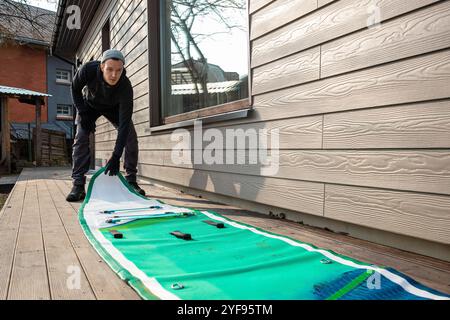 Homme pliant un stand-up paddleboard gonflable (SUP) sur une terrasse en bois à côté d'une maison moderne Banque D'Images