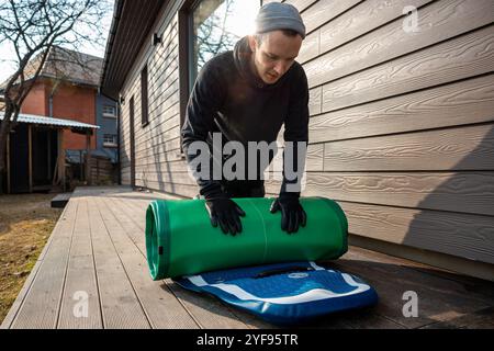 Homme pliant un stand-up paddleboard gonflable (SUP) sur une terrasse en bois à côté d'une maison moderne Banque D'Images