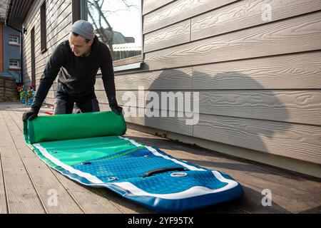 Homme pliant un stand-up paddleboard gonflable (SUP) sur une terrasse en bois à côté d'une maison moderne Banque D'Images