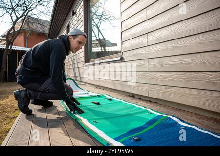 Homme pliant un stand-up paddleboard gonflable (SUP) sur une terrasse en bois à côté d'une maison moderne Banque D'Images