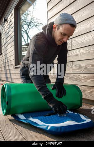 Homme pliant un stand-up paddleboard gonflable (SUP) sur une terrasse en bois à côté d'une maison moderne Banque D'Images