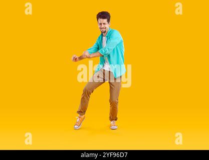 jeune brun guy danse porter chemise bleue, pantalon marron et baskets isolés sur fond jaune Banque D'Images