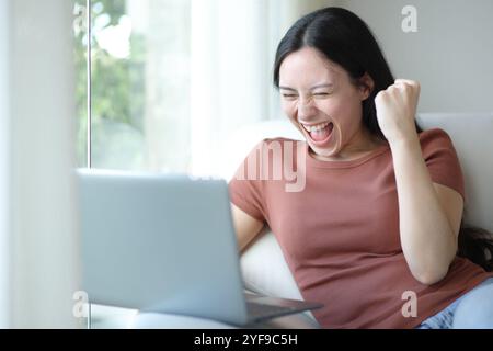 Femme asiatique excitée utilisant un ordinateur portable assis dans un canapé à la maison Banque D'Images