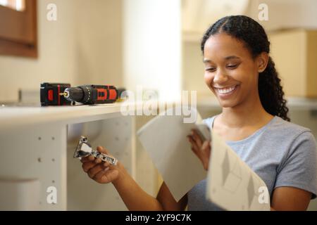 Femme noire heureuse lisant le manuel d'instructions installant des meubles de cuisine à la maison Banque D'Images