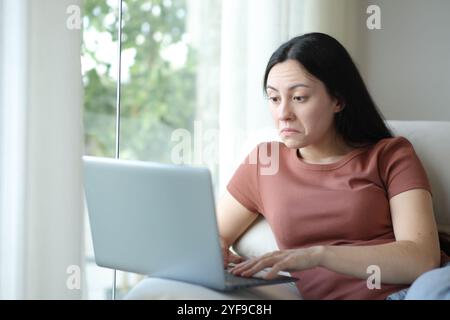 Femme asiatique perplexe utilisant un ordinateur portable assis sur un canapé à la maison Banque D'Images