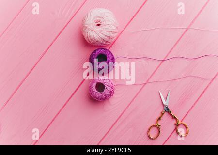 Un rouleau d'écheveaux roses et violets de fil et de ciseaux en fer repose sur une surface rose en bois, parfait pour la décoration, l'emballage cadeau et la couture. Banque D'Images